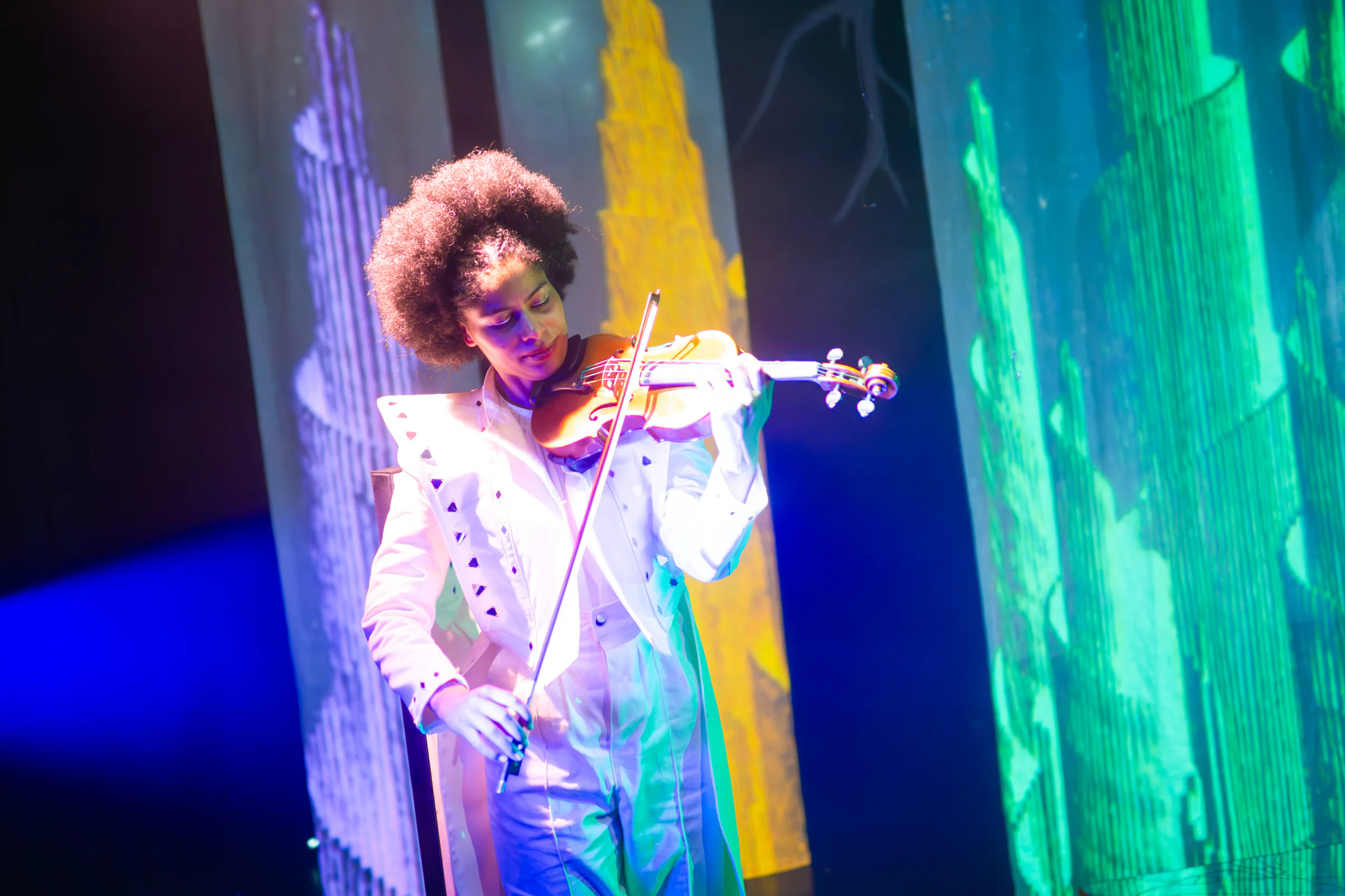 Fay Guiffo plays violin beside luminous green and blue scenic panels.